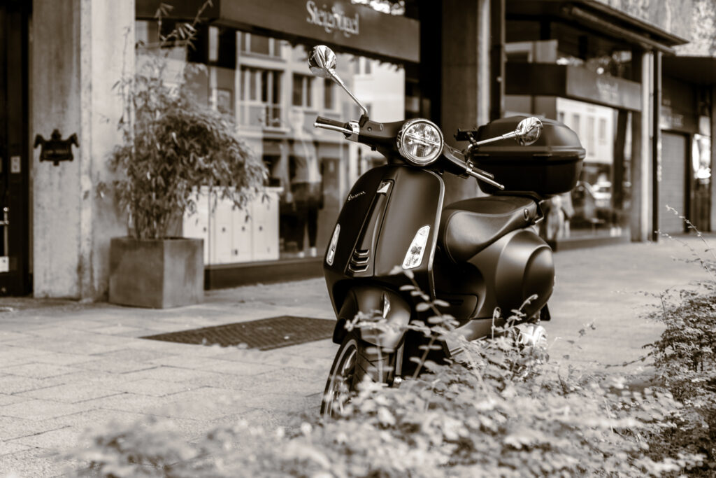 Eine schwarze Vespa Primavera hinter einer kleinen Hecke